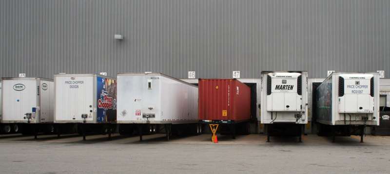 FIGURE 4. Trailer-Type Equipment: Dry Vans, an Ocean Container, and Refrigerated Trailers
This image shows six semi-trailers positioned at a row of warehouse loading bays. Three of the semi-trailers are conventional dry vans, one is a container used in ocean shipping, and two have refrigerator units attached.
FIGURE 4. Trailer-Type Equipment: Dry Vans, an Ocean Container, and Refrigerated Trailers
This image shows six semi-trailers positioned at a row of warehouse loading bays. Three of the semi-trailers are conventional dry vans, one is a container used in ocean shipping, and two have refrigerator units attached.